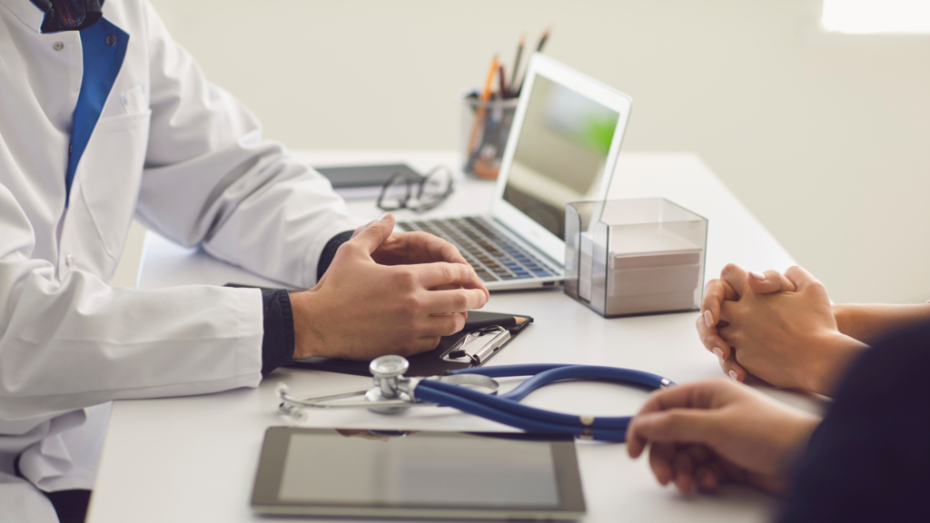 A close-up of a doctor wearing a white coat sitting at a desk, with hands clasped in discussion with a patient. A stethoscope, clipboard, and tablet are on the desk, alongside an open laptop and office supplies. The setting suggests a professional medical consultation.