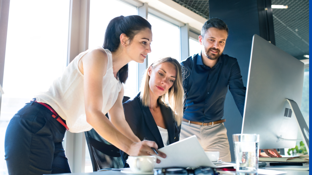 A team of three business professionals engaged in a discussion during a meeting. One woman is pointing at a document while another woman and a man attentively review it, with a large computer screen in the background. The modern office setting features natural light and a collaborative atmosphere.