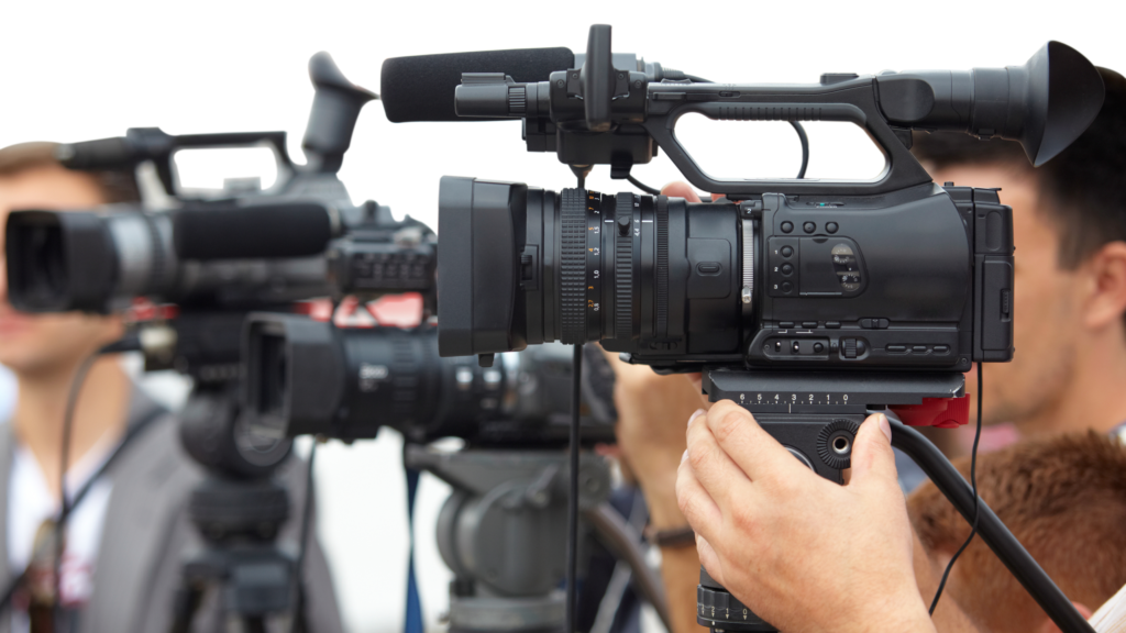 A close-up view of professional video cameras being operated during a media event. The image shows multiple cameras with attached microphones, capturing the scene, with blurred figures of camera operators in the background. It highlights the field of videography and media production.